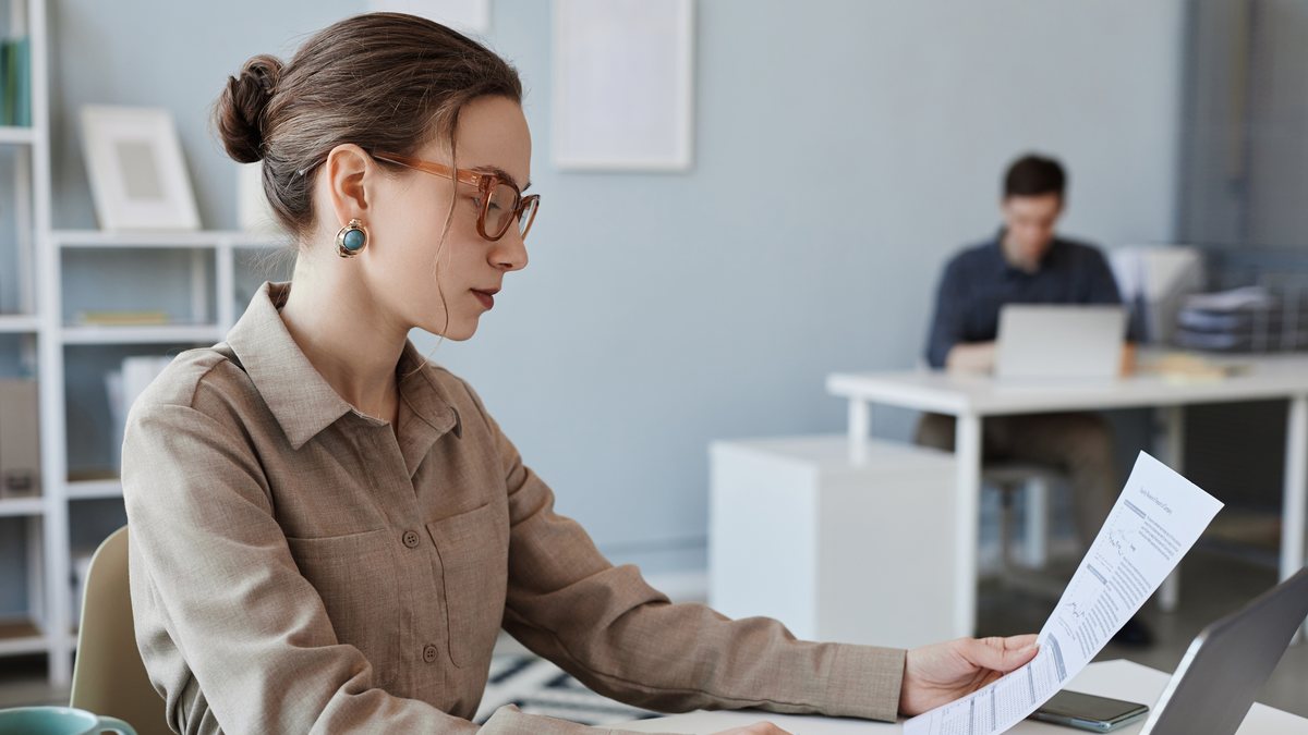 A business owner in glasses reviewing a printed document at her desk in a bright office