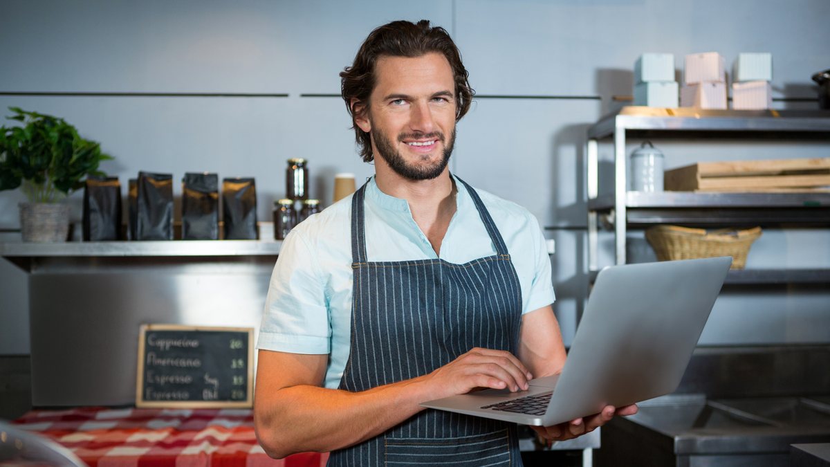 A cafe owner in an apron smiling while working on a laptop behind the counter