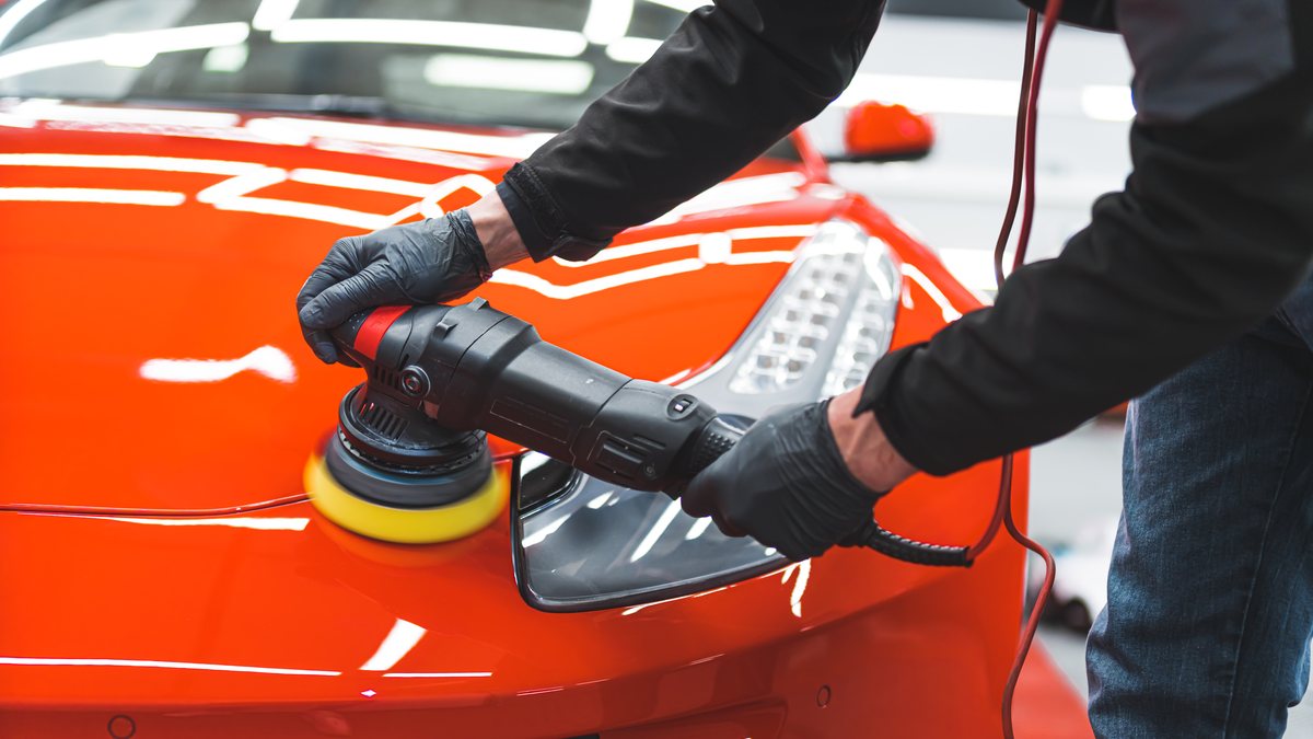 A detailer in black gloves machine-polishing the bonnet of a bright orange car