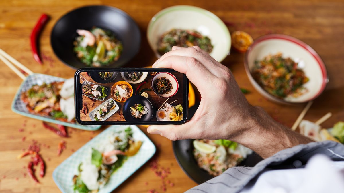 A hand holding a smartphone to photograph a table of freshly served dishes from above